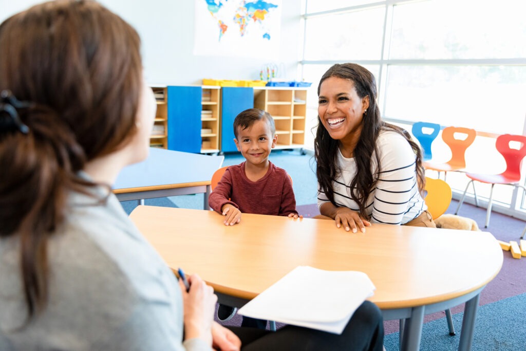 The Benefits of Early Childhood Education: A Guide for Parents 2 The cute preschool age boy watches quietly as his mid adult mom and his unrecognizable female teacher smile and laugh during the parent teacher conference.