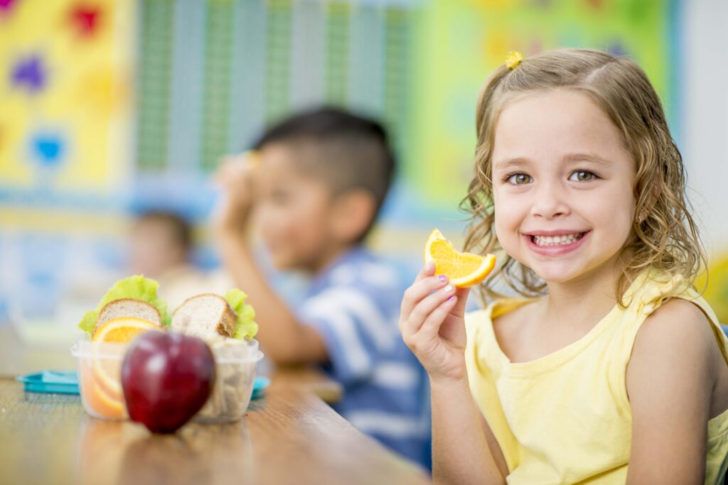 Healthy Snack Ideas for Kids: Nutritious and Delicious 10 A young girl is sitting indoors in her elementary school classroom. She is smiling at the camera while eating an orange.